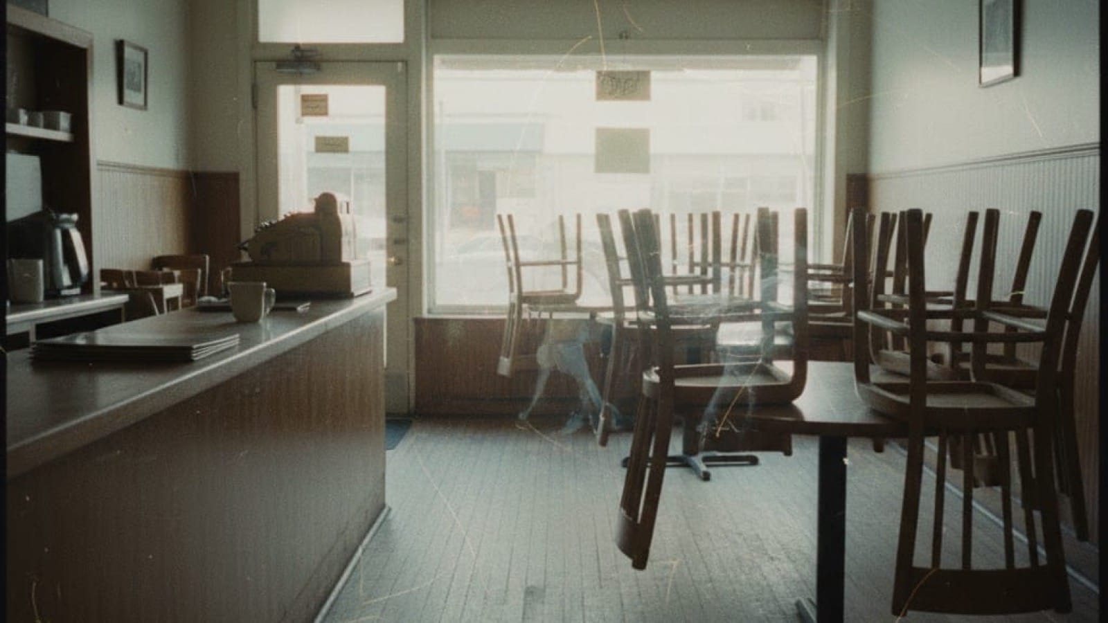 Empty family restaurant at dawn. Chairs stacked on tables, morning light through windows. Ghostly figures of future customers drift through the space like smoke.