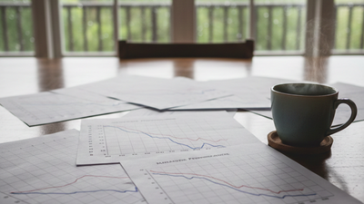 Desk in natural, overcast Hawaiian light. Scattered papers show declining performance graphs. A steaming coffee cup sits nearby, with rain visible through a large window. 