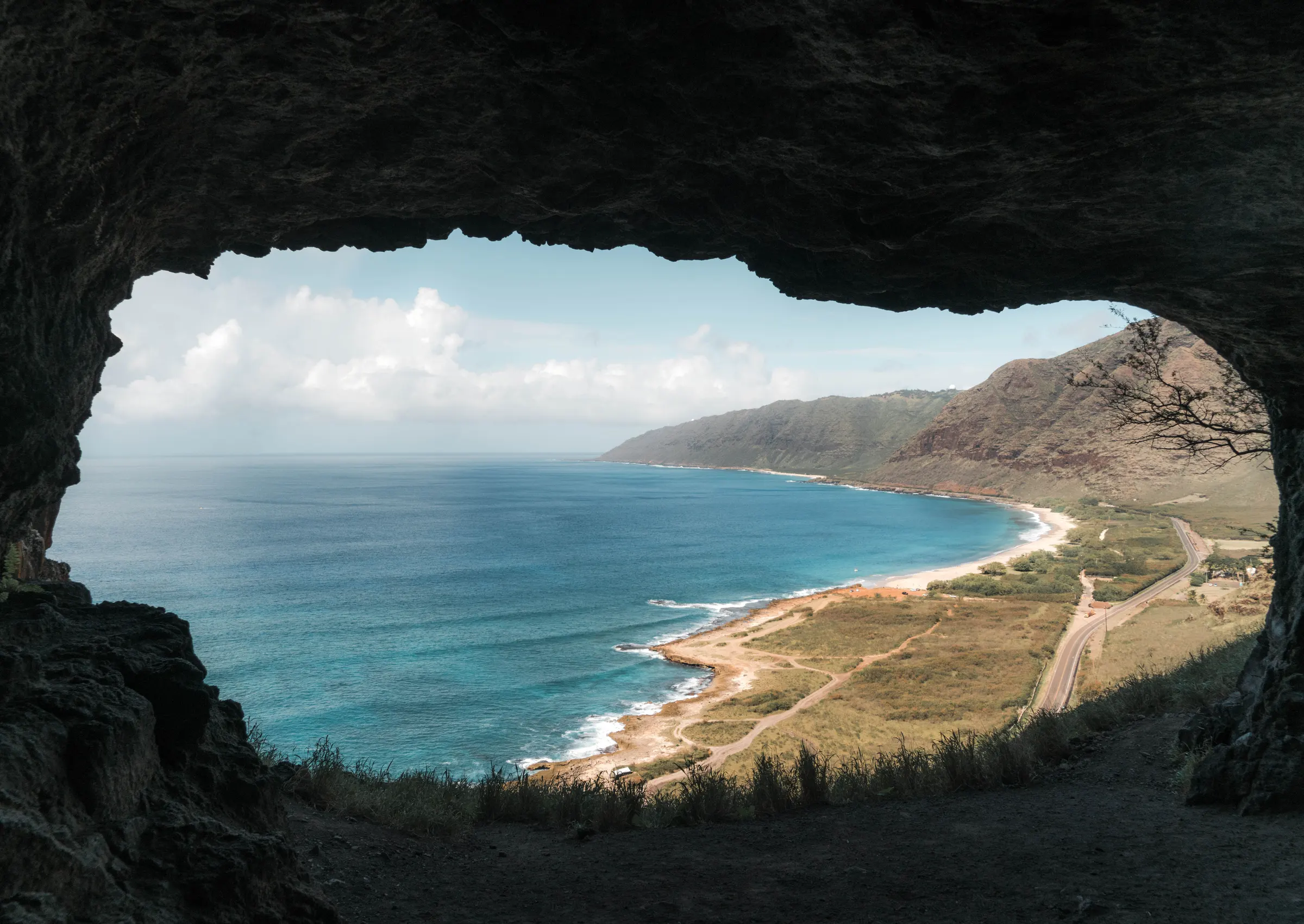 A Hawaii shoreline seen from inside a sea cave: surf meeting a sand beach below green ridges